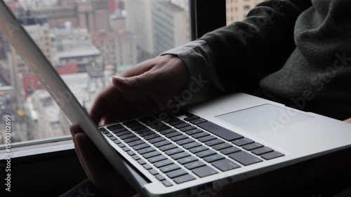 Handheld close-up of fingers typing on a laptop by a window overlooking Brooklyn and Manhattan below  and in the background. 