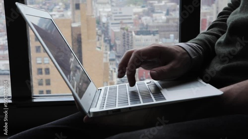 Handheld close-up of fingers typing on a laptop by a window overlooking Brooklyn and Manhattan below  and in the background. 