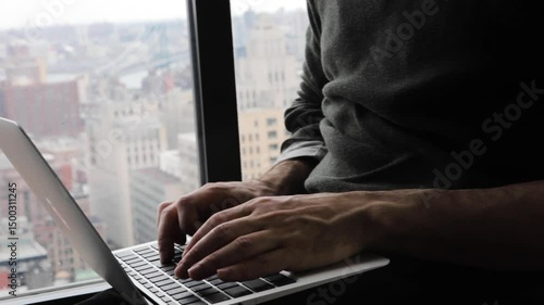 Handheld close-up of fingers typing on a laptop by a window overlooking Brooklyn and Manhattan below  and in the background. 