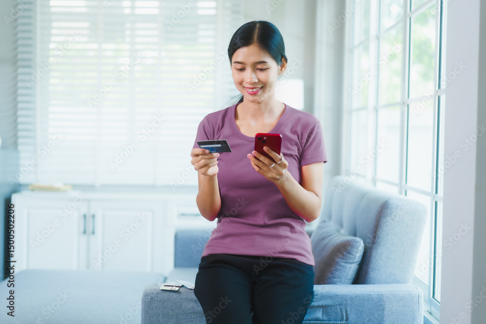 Fototapeta premium Happy Asian woman sits comfortably on a sofa,smiling while holding multiple credit cards,representing convenient online shopping,smart financial planning,modern digital consumption in a relaxed home