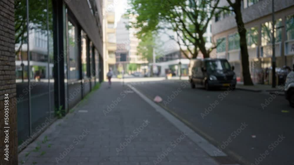 Defocused view of a european city street with a blurred woman walking on a sidewalk surrounded by modern buildings and green trees under soft daylight. Defocused view of a european city street with a blurred woman walking on a sidewalk surrounded by modern buildings and green trees under soft daylight.