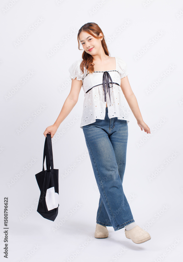 Asian young beautiful woman cheerful confident long curly hair wearing white puff sleeve blouse black ribbon blue jeans holding black canvas shopping bag look at camera stand pose on white background. Asian young beautiful woman cheerful confident long curly hair wearing white puff sleeve blouse black ribbon blue jeans holding black canvas shopping bag look at camera stand pose on white background.