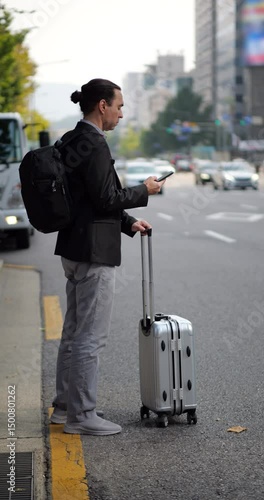 Urban lifestyle shot of businessman standing on road side, using smartphone while waiting for ride with his trolley bag by his side. Full length vertical shot with blurred background