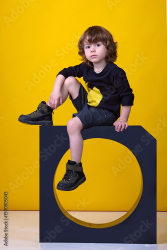 Little cute boy in black T-shirt, black shorts and sneakers posing sitting in studio on yellow background