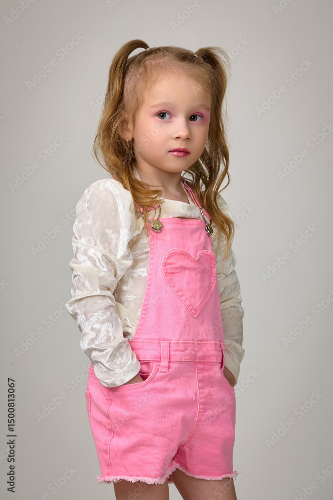 Little cute girl in pink denim overalls and white blouse posing in studio on white background Little cute girl in pink denim overalls and white blouse posing in studio on white background