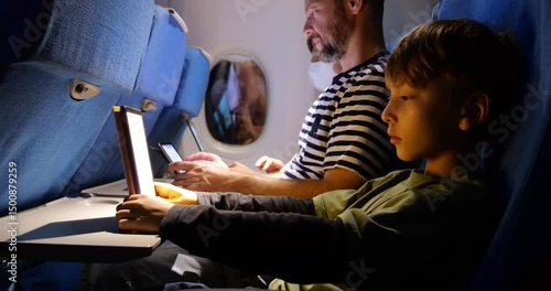 Nighttime airplane interior showing family during flight. Young boy reading on tablet or e-book device, while father uses tablet and mother works on laptop in background