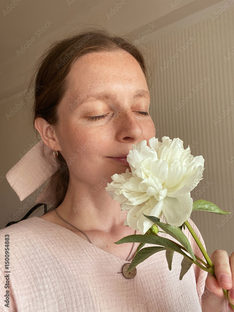 Young elegant girl smelling white peony flower with closed eyes in room Young elegant girl smelling white peony flower with closed eyes in room