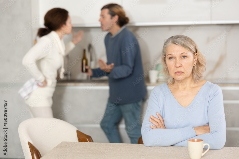 Fototapeta premium Mature woman is sitting in kitchen and is upset by quarrel between her son and daughter-in-law. Wife screams and insults her husband