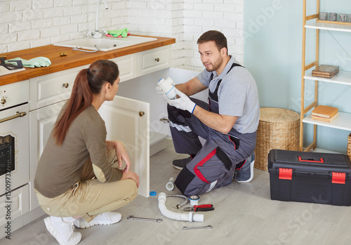 Bild auf Leinwand Professional male plumber trying to fix siphone in kitchen sink, explaining woman client what is wrong, trying to solve problem with pipe, cleaning out the clogged drain