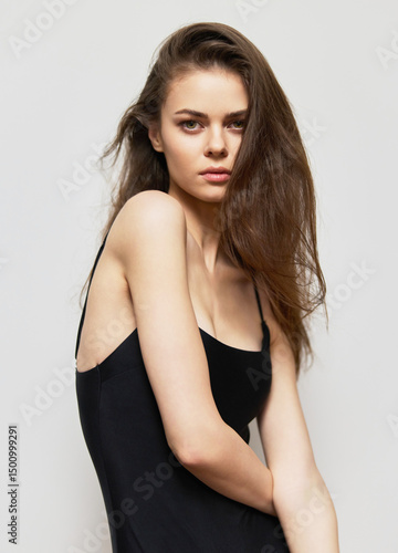 Confident young woman with long hair posing in minimalistic black outfit against a soft gray background, expressing elegance and natural beauty.