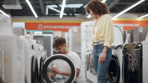 Couple shopping for washing machine, partner highlighting key features while discussing options in electronics store, capturing decision-making process in slow motion