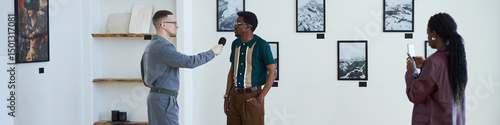 Fototapet Header with black man standing in an art gallery, being interviewed by a man holding microphone while woman taking notes is nearby