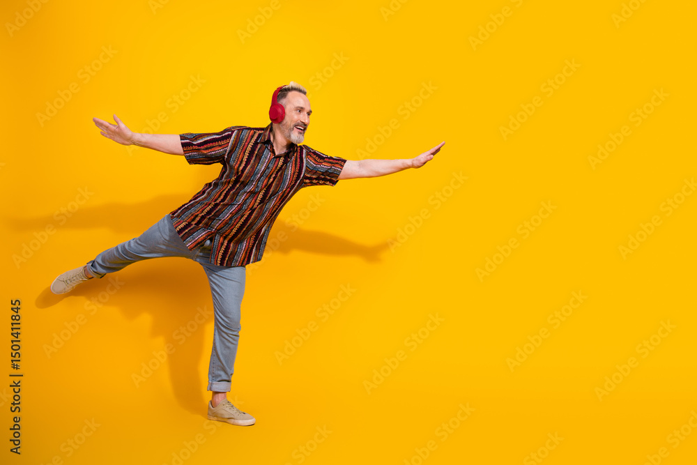 Middle aged Man in Vibrant Shirt Balances Playfully Against Yellow Background Listening to Headphones Middle aged Man in Vibrant Shirt Balances Playfully Against Yellow Background Listening to Headphones