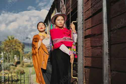 Two indigenous ecuadorian models are posing near an old wooden train, wearing colorful traditional andean clothing and accessories