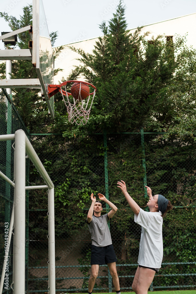 Fototapeta premium Young man and woman playing basketball on an outdoor court in a residential yard. Sunny summer day, green trees, clear blue sky. Active lifestyle and urban sport