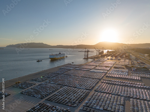 RORO Cargo Ship and New Cars on Large Full Parking Lot at Sunset in Port Ready for Distribution. Car Storage, Automotive Industry. Ocean and Mountains. Setubal, Portugal. Aerial View