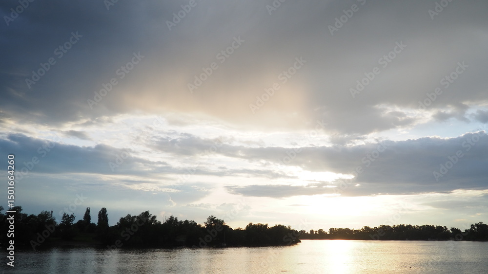 Obraz premium Witness the breathtaking spectacle of clouds as they dance across the sky, casting shadows over calm waters at sunset. Natures palette transitions with stunning hues of orange and blue