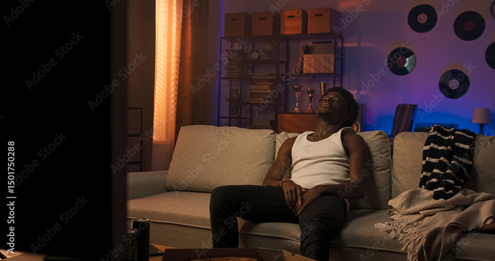 Obraz premium A sporty male with a multi-racial background sits on a couch in a white strapped T-shirt, looking away in silence, expressing deep thought and subtle disappointment.