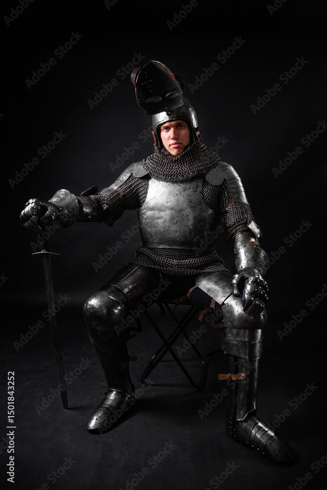 Naklejka premium Portrait of a knight in full plate armor holding a sword posing sitting on chair isolated on a dark background.