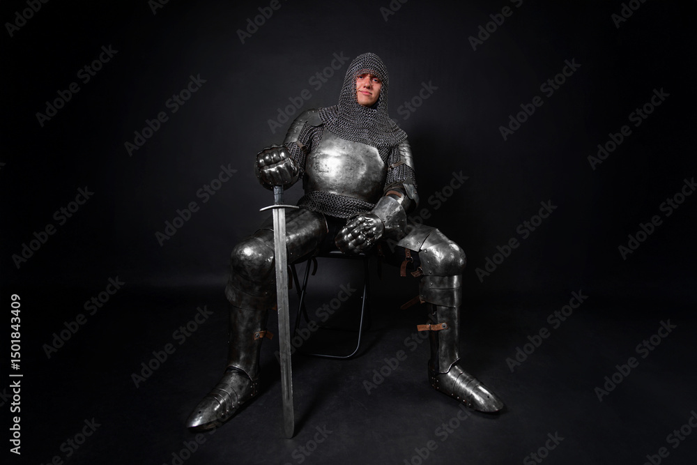 Naklejka premium Portrait of a knight in full plate armor holding a sword posing sitting on chair isolated on a dark background.