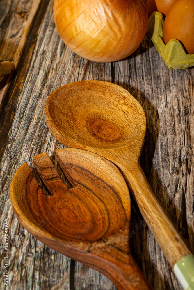Rustic kitchen still life featuring wooden salad servers, a whole onion, and a carton of brown eggs, set against a weathered wooden surface. Rustic kitchen still life featuring wooden salad servers, a whole onion, and a carton of brown eggs, set against a weathered wooden surface.