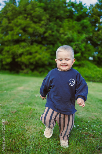 Toddler laughing and enjoying warm sunny day. Happy 2 year old boy in t-shirt and shorts running on green grass and laughing outdoors. Child playing on public playground on sunny summer day.