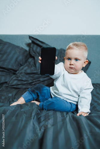 Handsome toddler boy in a white t-shirt and jeans sits on the bed and watches cartoons on the phone. 2 year old boy plays with a mobile gadget. Modern digitalization of toddlers.