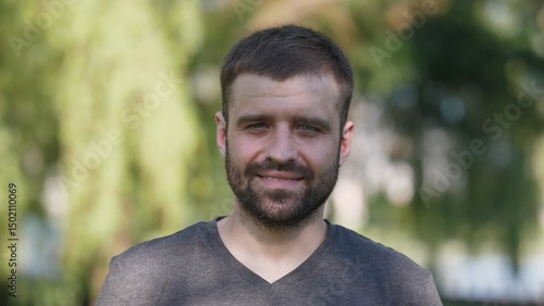 A man with a short beard stands confidently in a park, enjoying a bright sunny afternoon while surrounded by lush greenery, radiating positivity and warmth.