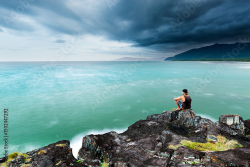 Contemplative young man sitting on a rock, looking at stormy ocean seascape and tropical coastline at Cape Tribulation, Queensland, Australia