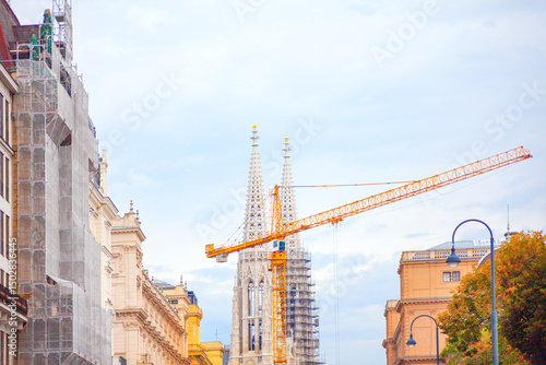 Vienna Votivkirche, a neo-Gothic church with twin spires, under construction with a yellow crane looming overhead. Surrounding buildings, some with scaffolding, frame scene under a cloudy sky