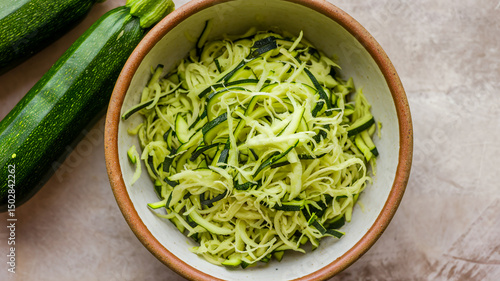 A bowl filled with finely grated green vegetables, likely zucchini, placed on a textured surface. Adjacent to the bowl are two whole zucchinis with their green hues intact