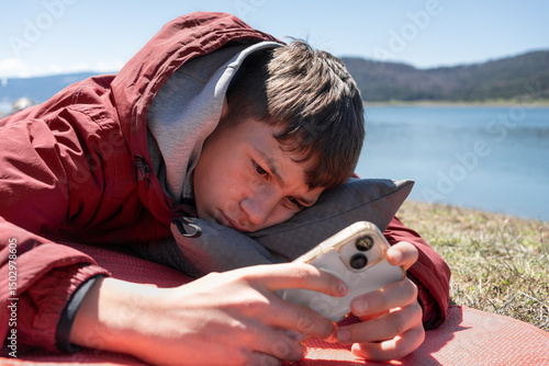 Bored teen scrolling phone by Lake, ignoring nature. Close-up of bored teenager lying near scenic lake, scrolling phone with frown. Emotionally distant, lost in technology and unaware of beauty around