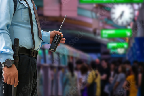 Close-up of a uniformed security guard holding a walkie-talkie at a busy metro platform filled with commuters and signage.