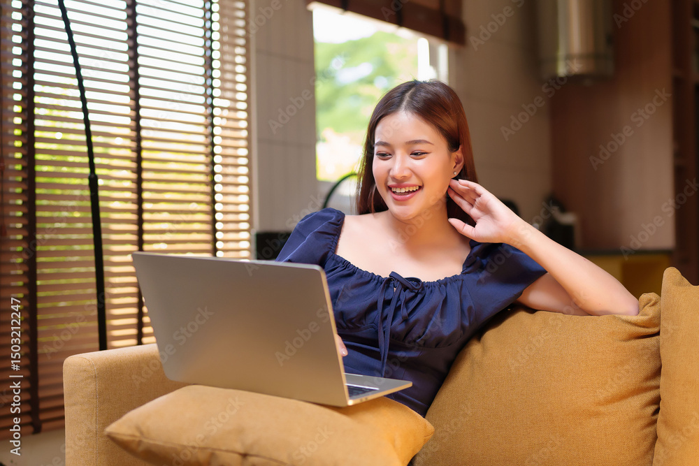 Naklejka premium Asian Woman Smiling While Using Laptop on Sofa at Home Asian young woman using computer for social media searching or communicating with friends