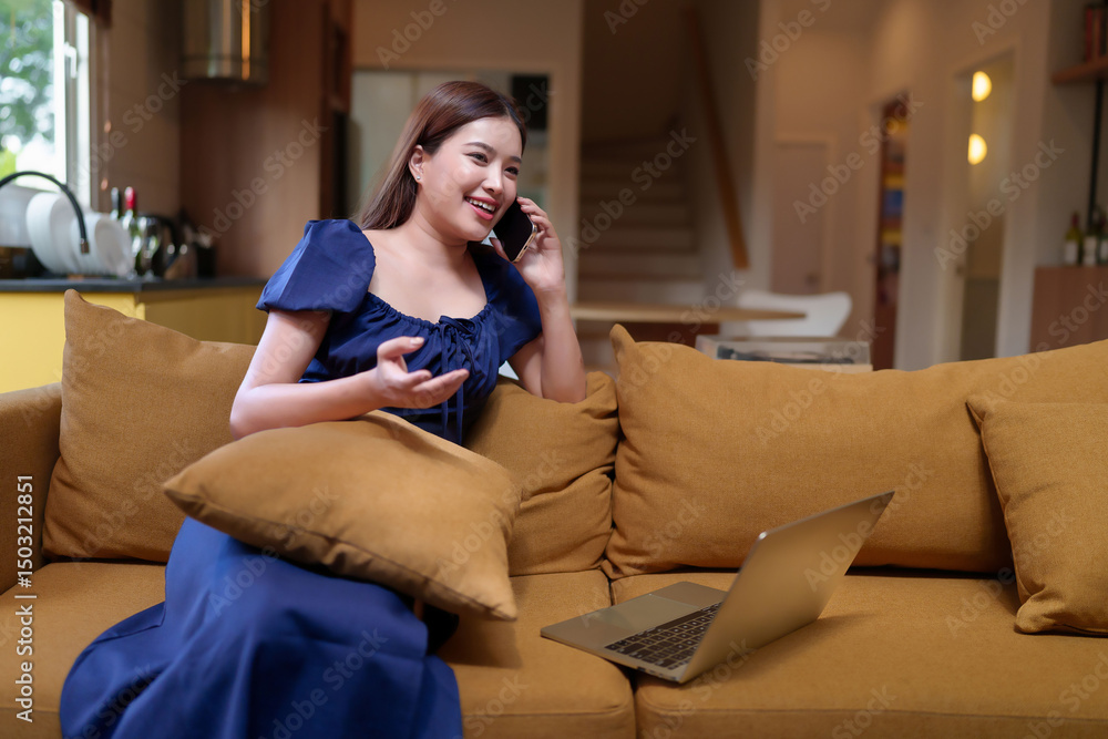 Obraz premium Happy Asian woman talking on her mobile phone while sitting with a laptop on a yellow sofa at home in a relaxed atmosphere