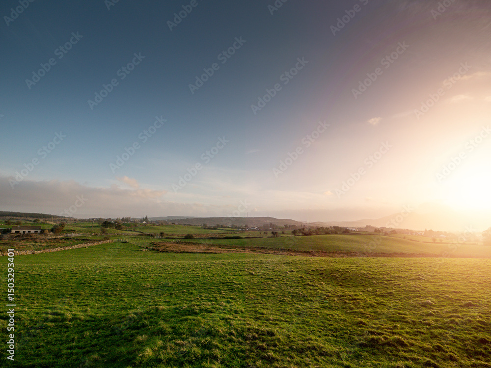 Obraz premium A large field of grass with a few houses in the distance. The sky is cloudy and the sun is setting. Calm and relaxing nature scene with rich color and warm summer vibe. Nobody. Picturesque view.