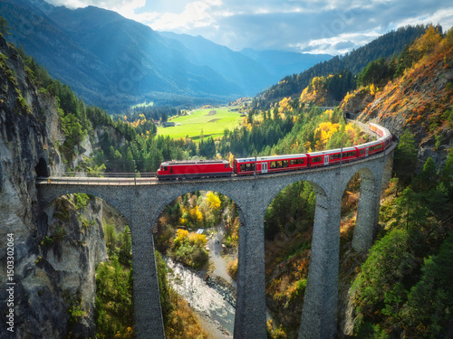 Aerial view of modern red train on Landwasser viaduct in alpine mountains, colorful forest at sunset in autumn. Bernina Express, Switzerland. Top view of train in Alps, railroad, green trees in fall