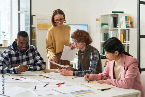 Group of diverse students learning new languages by reading, discussing and writing at shared table in a modern classroom with reference materials