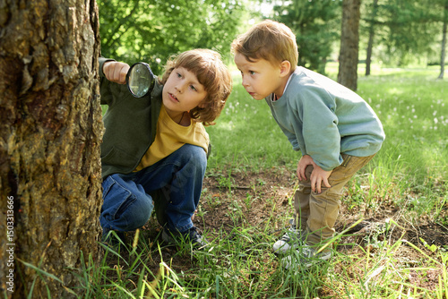 Full length portrait of two young brothers using magnifying glass and looking at bugs and insects on trees in green forest adventuring in nature together