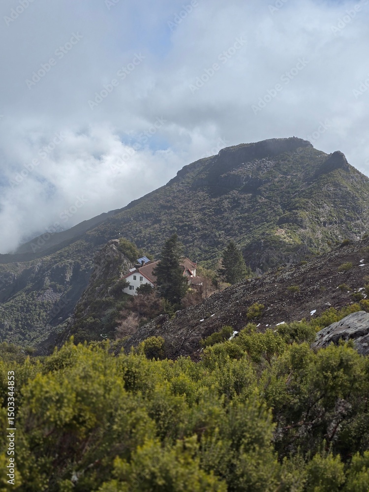 Fototapeta premium Clouds drift between the rugged peaks of Pico Ruivo, while a tiny house in the distance anchors the vast mountainous terrain.