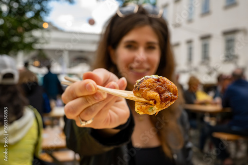 Young woman smiling while holding a takoyaki ball with chopsticks toward the camera. Close-up focus on the octopus snack in front of her.