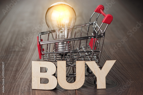 Photo of shopping trolley with glowing bulb in it and word buy made up of wooden letters. Buy smart concept on the wooden background.