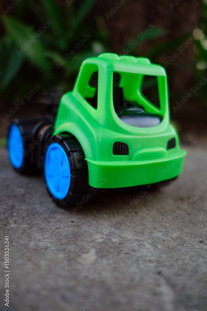 Fototapeta premium A vibrant green toy truck with blue wheels rests on a gravel surface Sunlight shines gently illuminating the toy against the backdrop of lush greenery creating a cheerful atmosphere