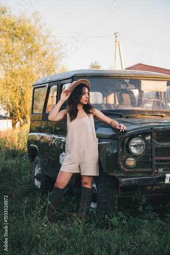 A beautiful woman in a light jumpsuit with a hat stands near a car in the village. Tinting