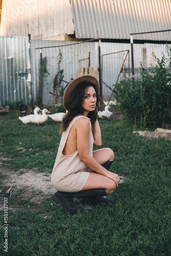 A beautiful woman in a light jumpsuit with a hat stands near a paddock with geese village. tinting
