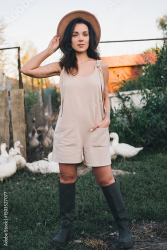 A beautiful woman in a light jumpsuit with a hat stands near a paddock with geese village. tinting