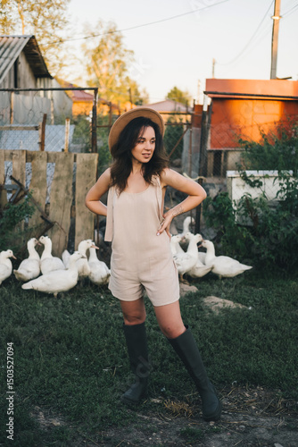A beautiful woman in a light jumpsuit with a hat stands near a paddock with geese village. tinting