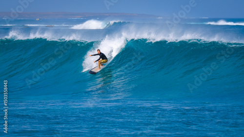 Male surfer in patterned yellow shorts is dropping into a large, clean wave under clear blue skies. Set wave is steep and hollow, rider is positioned low for control to make a proper bottom turn.