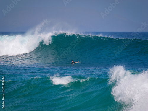 Surfer paddles out below a towering, curling wave about to break. Deep blue sea contrasts with foamy white crest, capturing the raw energy and thrill of ocean surfing in coastal waters of Lanzarote.