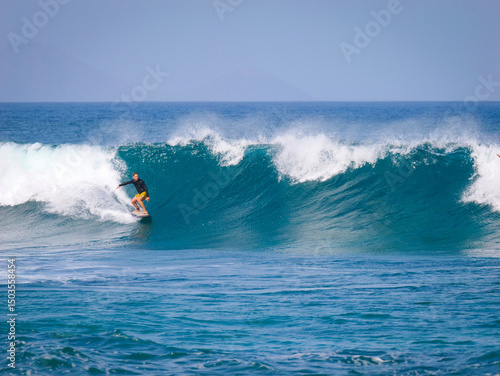 Male surfer in yellow shorts and a black rash guard charges on a large set wave with confidence. He leans into the carve as glassy wave crashes behind him. Big summer swell hit the Canary Islands.